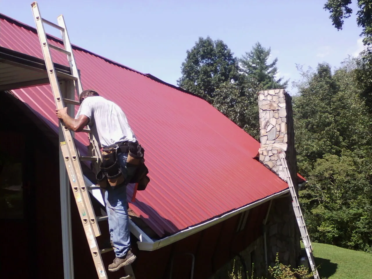 Roofer on a ladder beside a red metal roof and stone chimney — Soffit & Fascia Repair by Roofing Brothers in Riddle